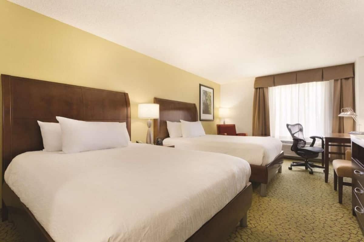 A hotel room with two beds, a desk, a chair, and a lamp.
