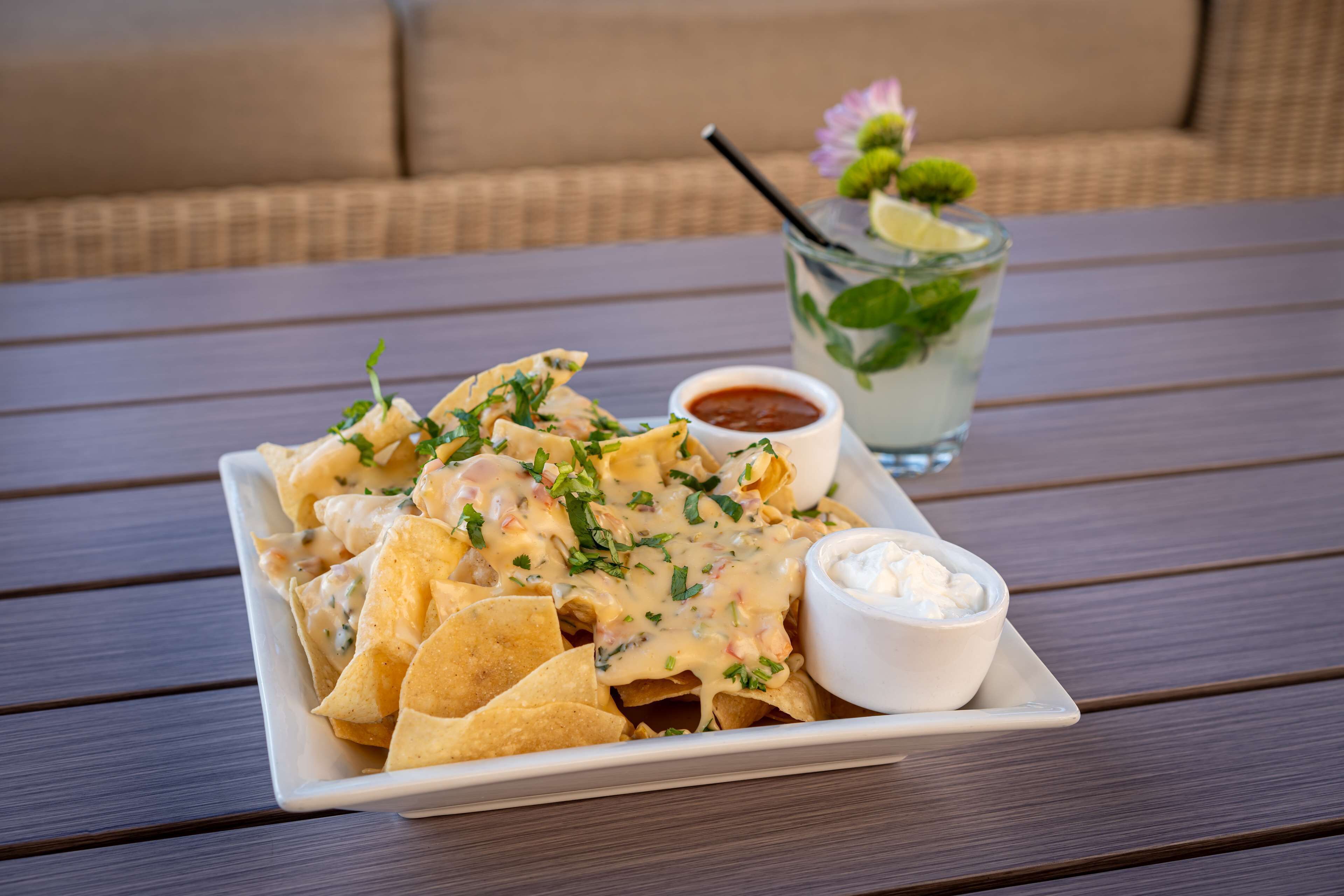 Un piatto di nachos con salsa cremosa e panna acida a parte, accompagnato da un bicchiere di tè freddo guarnito con menta.