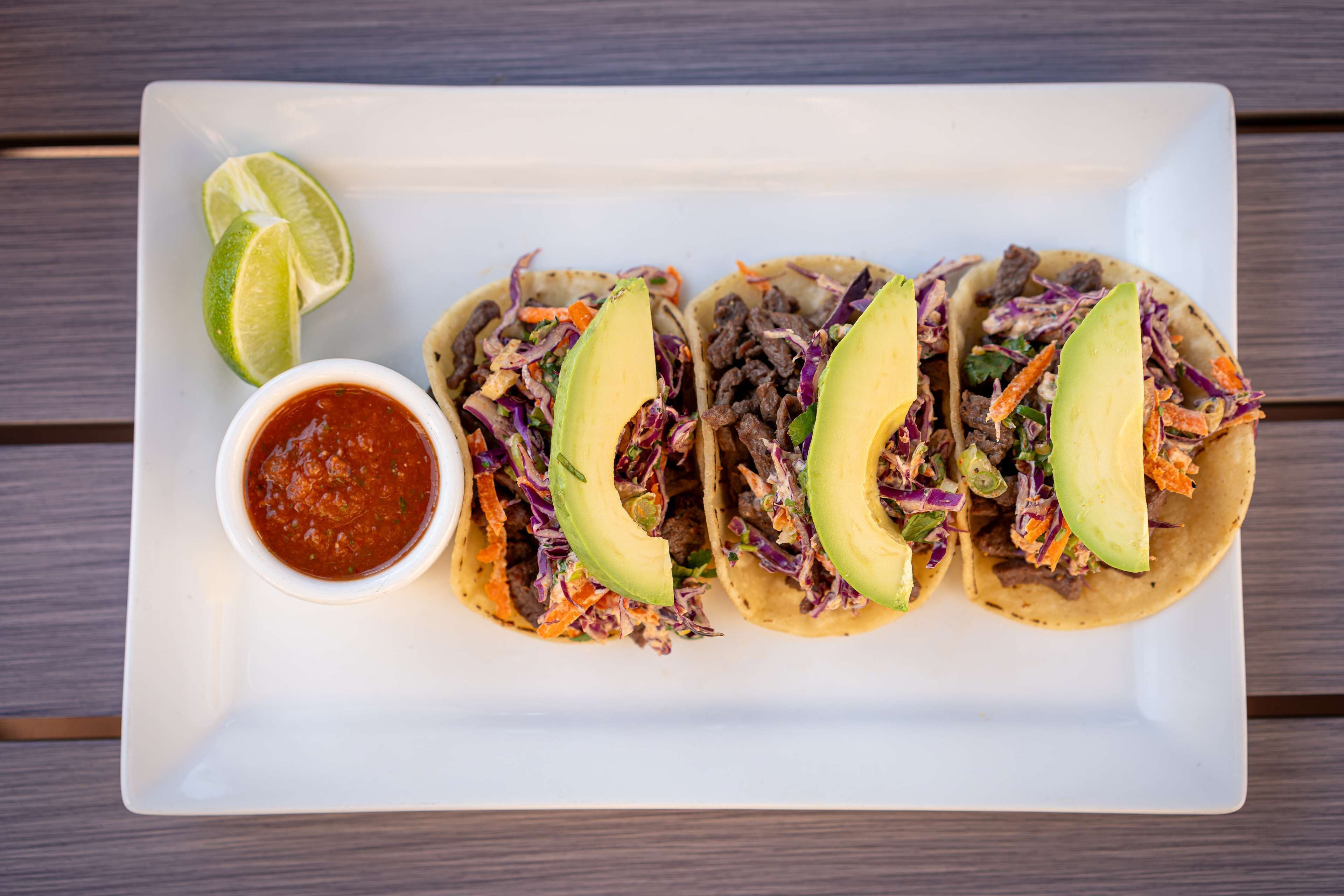 Three tacos with avocado and a small bowl of salsa on a white plate.