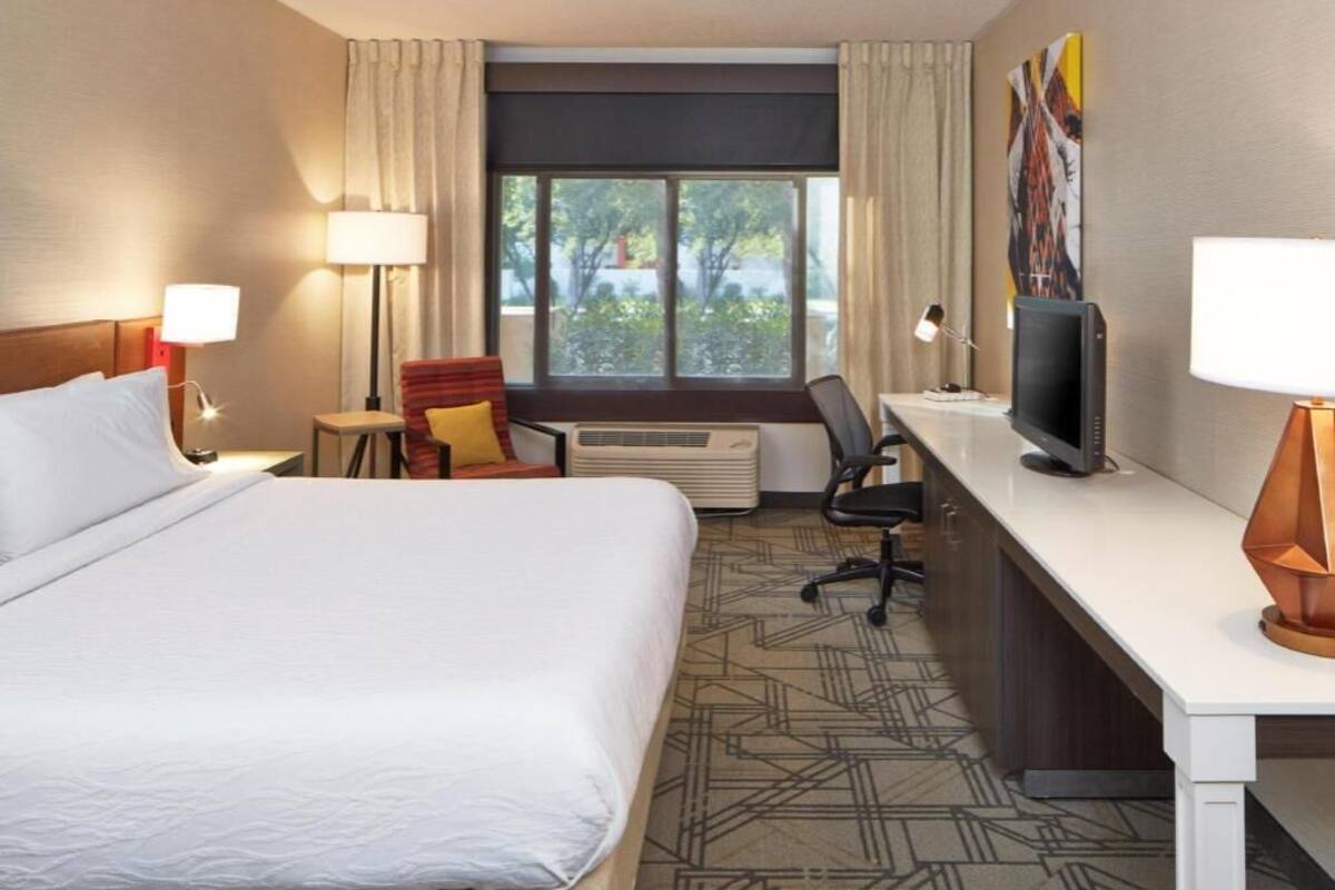 Ein Hotelzimmer mit Bett, Schreibtisch, Stuhl, Fernseher, Lampe und einem Fenster mit Vorhängen.