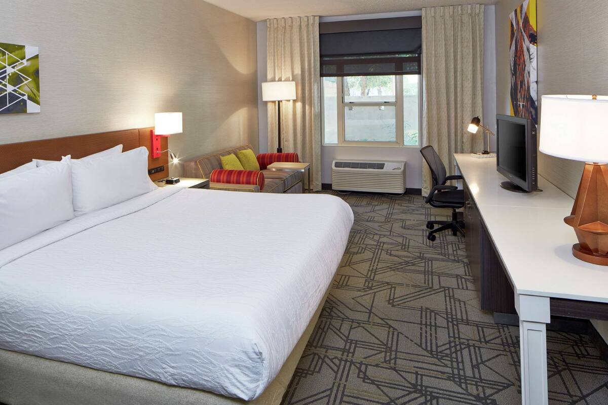 Ein Hotelzimmer mit einem großen Bett, einem Schreibtisch mit Computer, einem Sessel, einer Klimaanlage und einem Fenster mit Vorhängen.