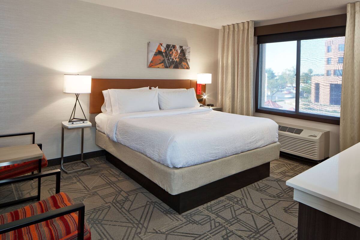 Ein Hotelzimmer mit einem großen Bett, einem Schreibtisch, einem Stuhl, einer Stehlampe und einem Fenster mit Blick auf Gebäude.
