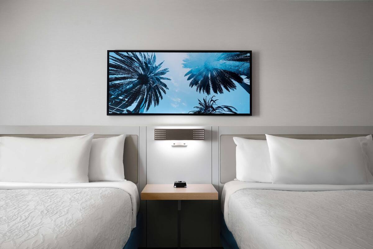 A hotel room with two beds, a nightstand, a wall-mounted light, and a framed picture of palm trees.