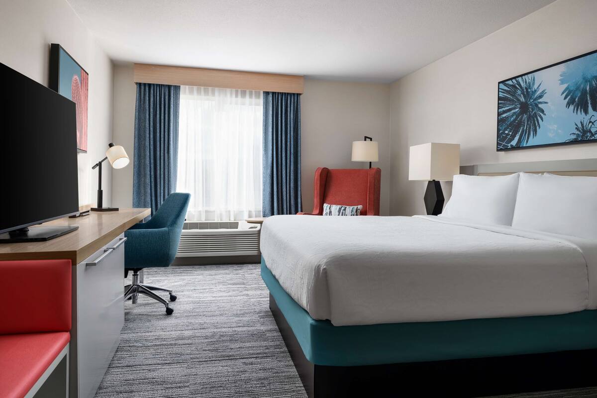 A hotel room with a bed, a desk with a TV, a chair, a desk lamp, a window with curtains, and a wall-mounted lamp.