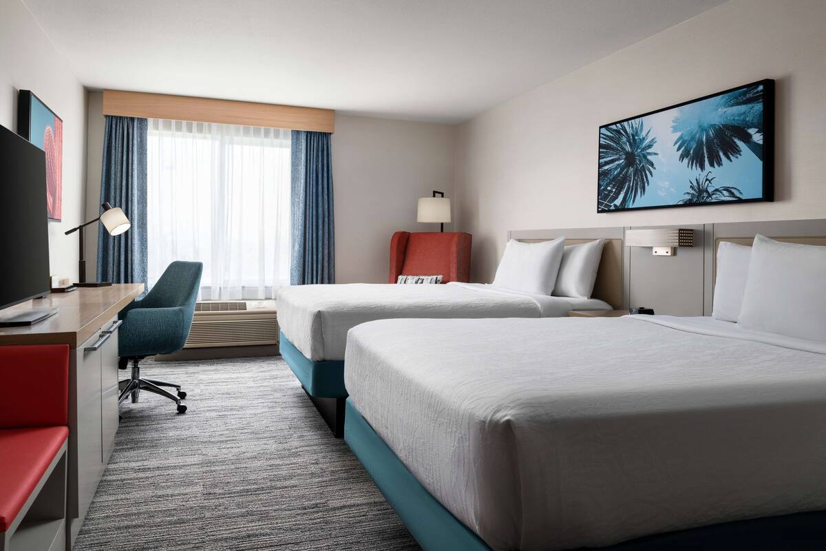 A hotel room with two beds, a desk with a chair, a TV, and a window with curtains.