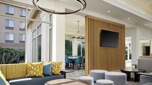 A modern hotel lobby with a sofa, ottomans, a round table, and a TV mounted on a wooden panel. - Hilton Garden Inn San Jose/Milpitas (Milpitas)