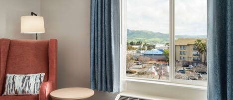 A hotel room with a red armchair, a small round table, a lamp, and a window with a view of a cityscape and mountains.