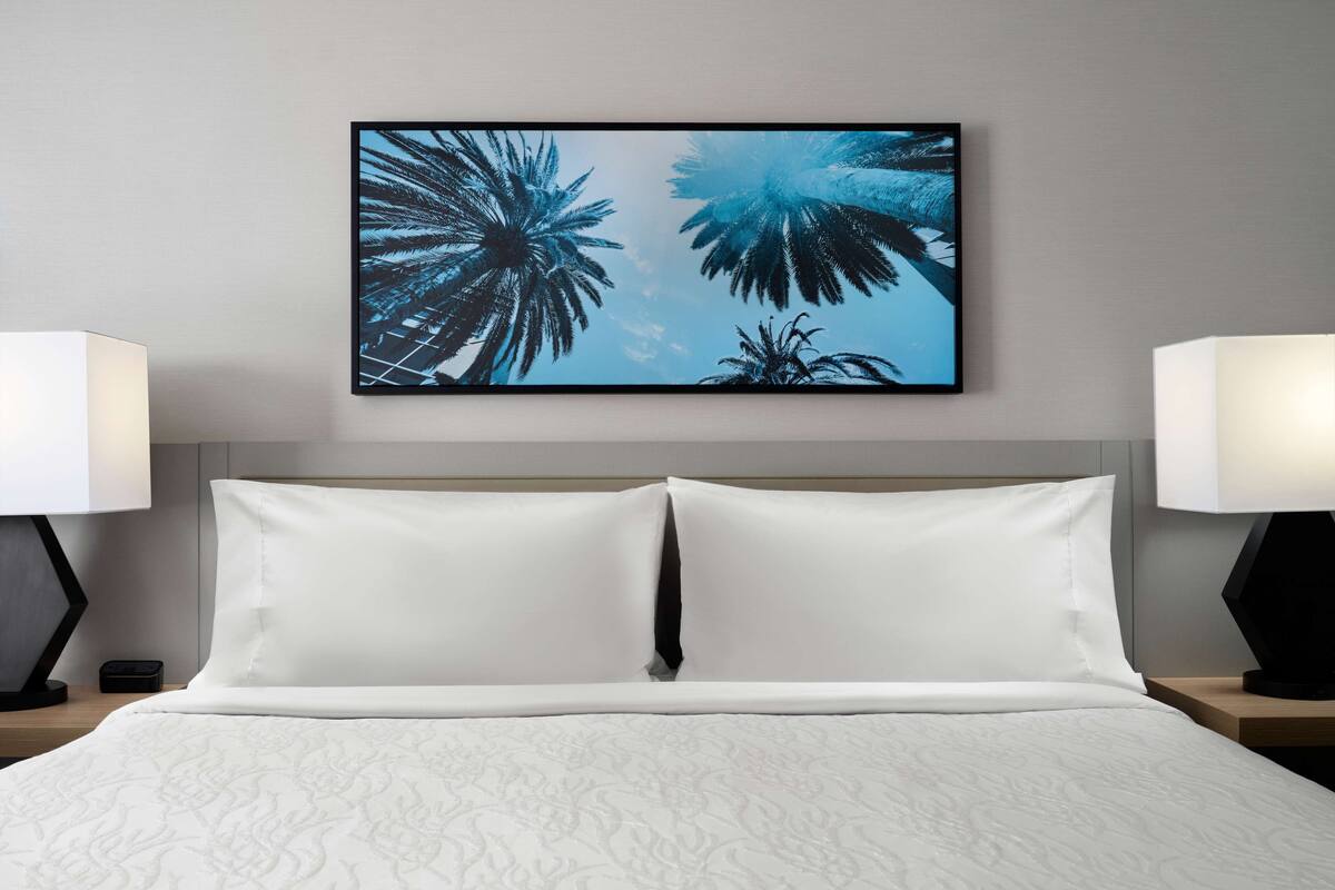 A neatly made bed with white linens and a large framed picture of palm trees on the wall above.