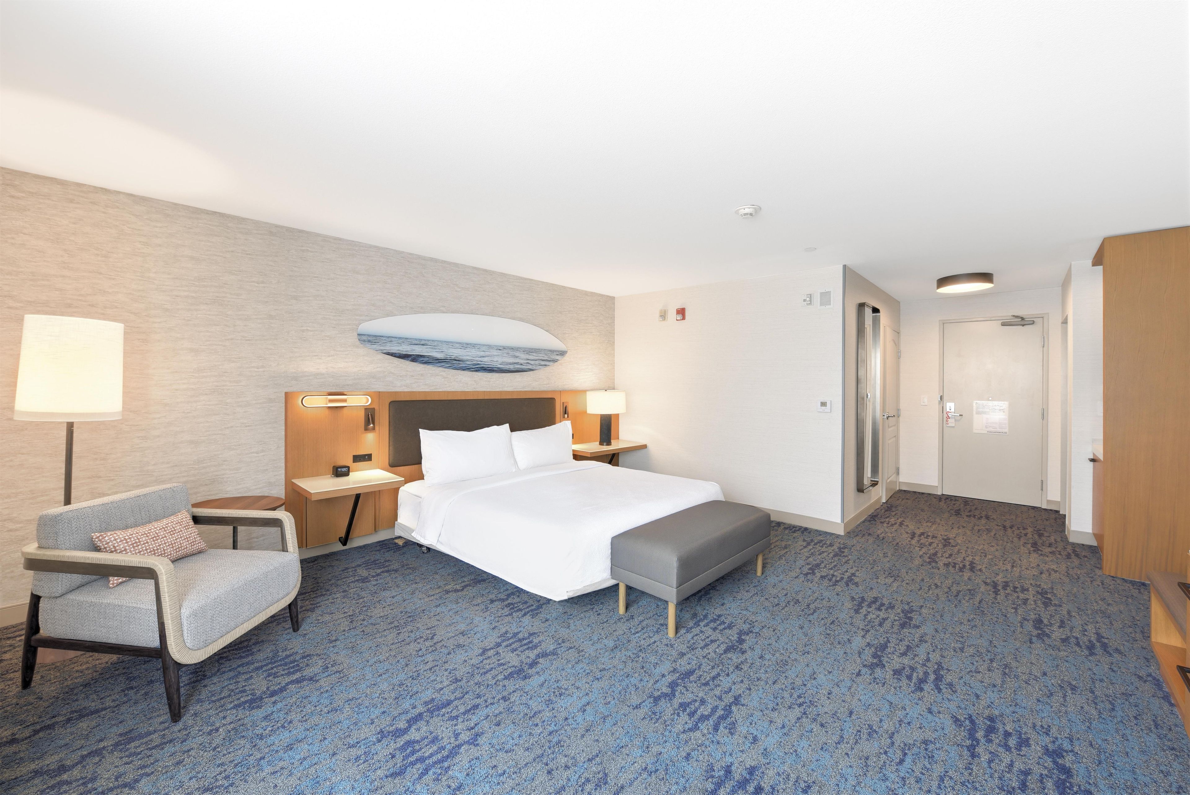 A hotel room with a bed, bedside tables, a desk, a chair, a bench, a wall-mounted artwork, and a door.