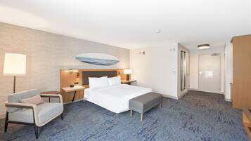 A hotel room with a bed, bedside tables, a desk, a chair, a bench, a wall-mounted artwork, and a door.