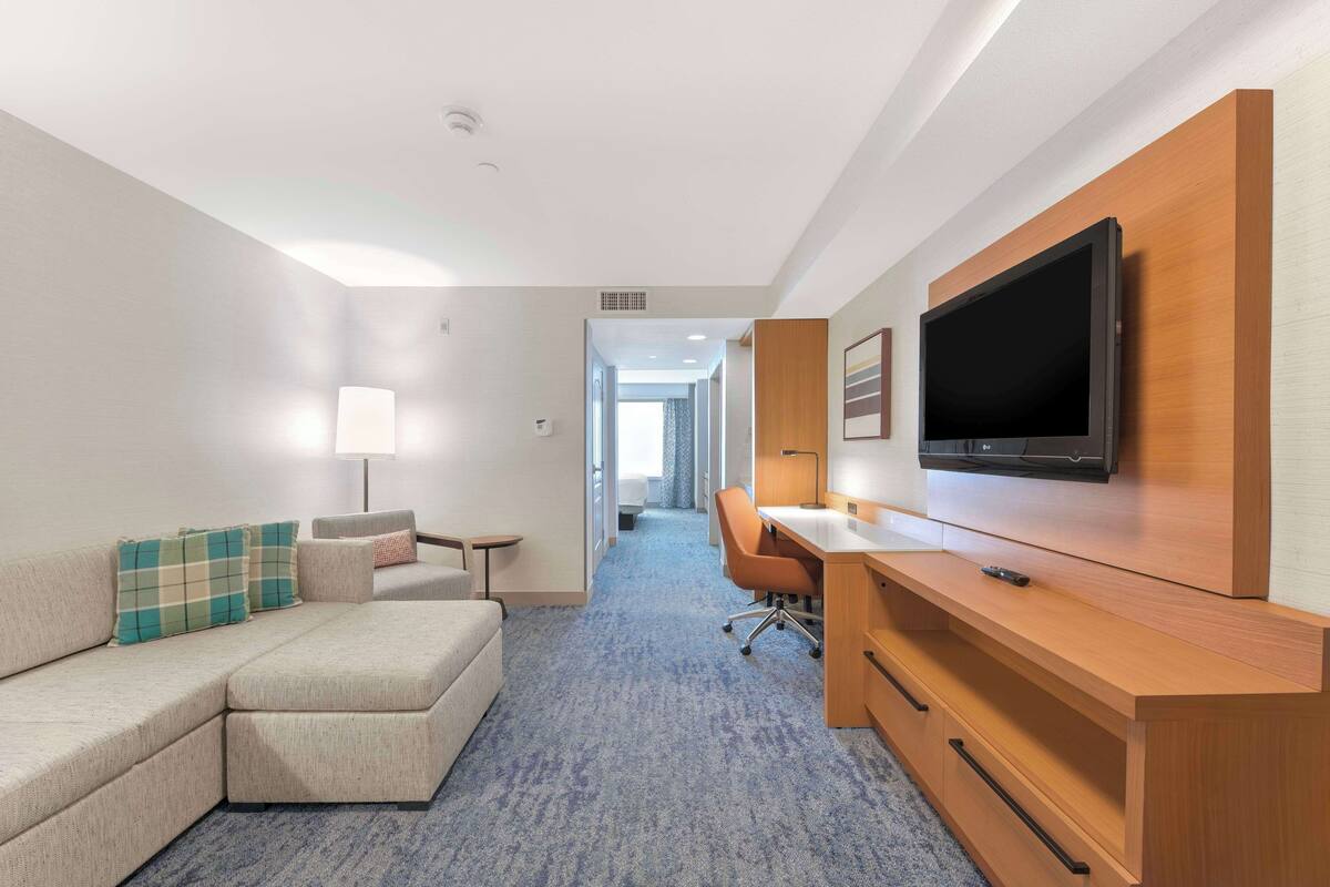 A hotel room with a sectional sofa, a desk with a flat-screen TV, and a lamp.