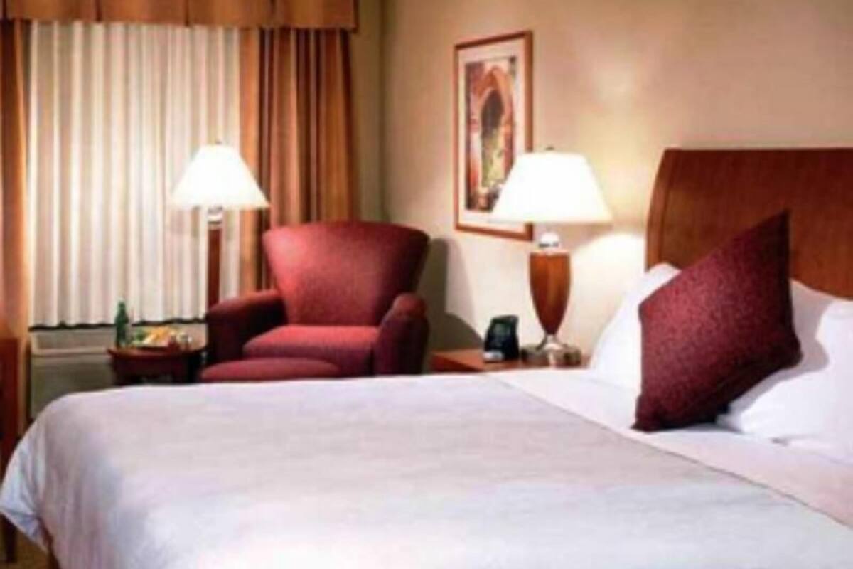 A hotel room with a bed, two bedside lamps, a red armchair, and a painting on the wall.