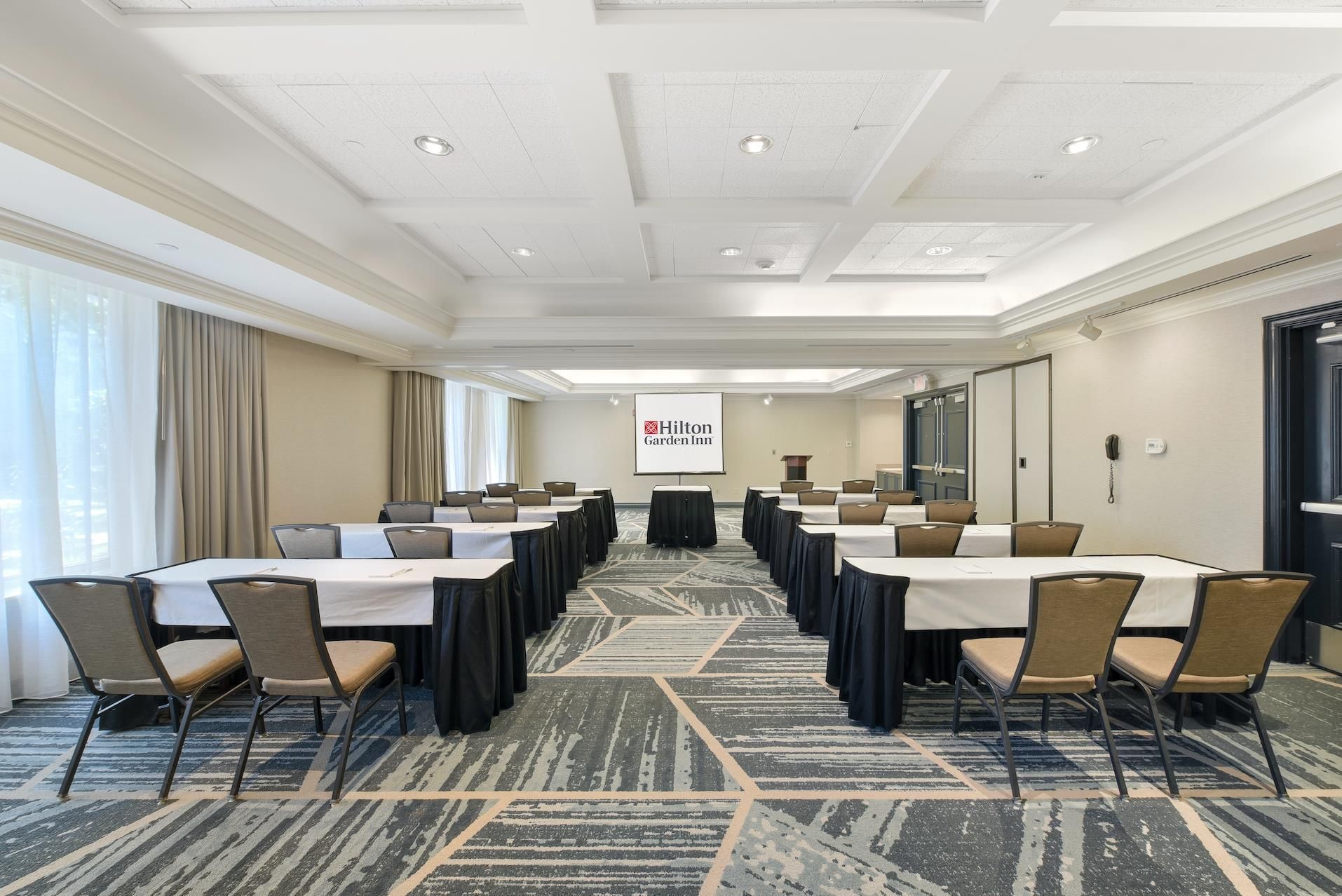 A conference room with multiple tables and chairs, a projector screen with the Hilton Garden Inn logo, and a carpeted floor.