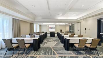 A conference room with multiple tables and chairs, a projector screen with the Hilton Garden Inn logo, and a carpeted floor.