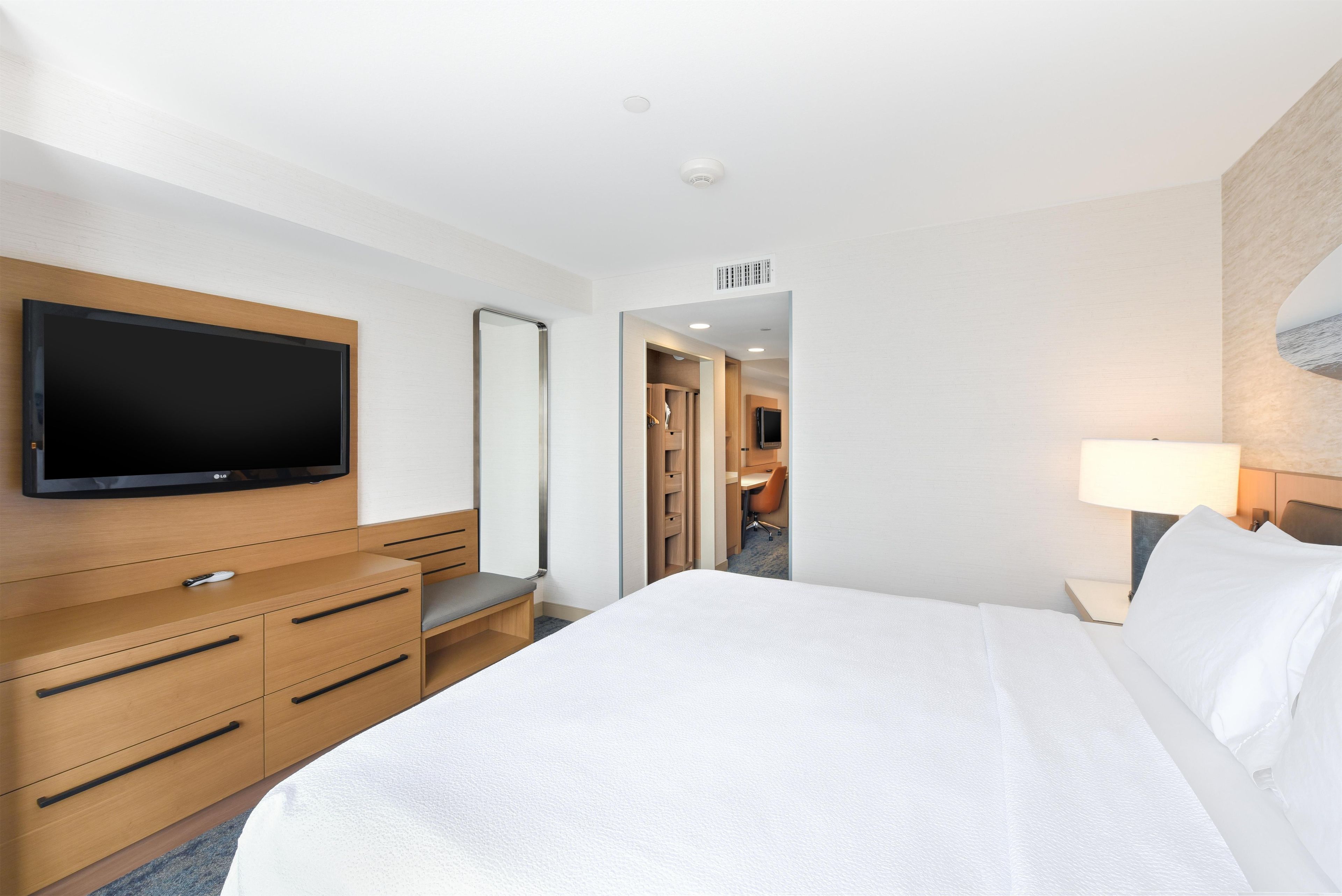 A hotel room with a bed, a TV mounted on the wall, a desk with a chair, and a bathroom visible through an open door.