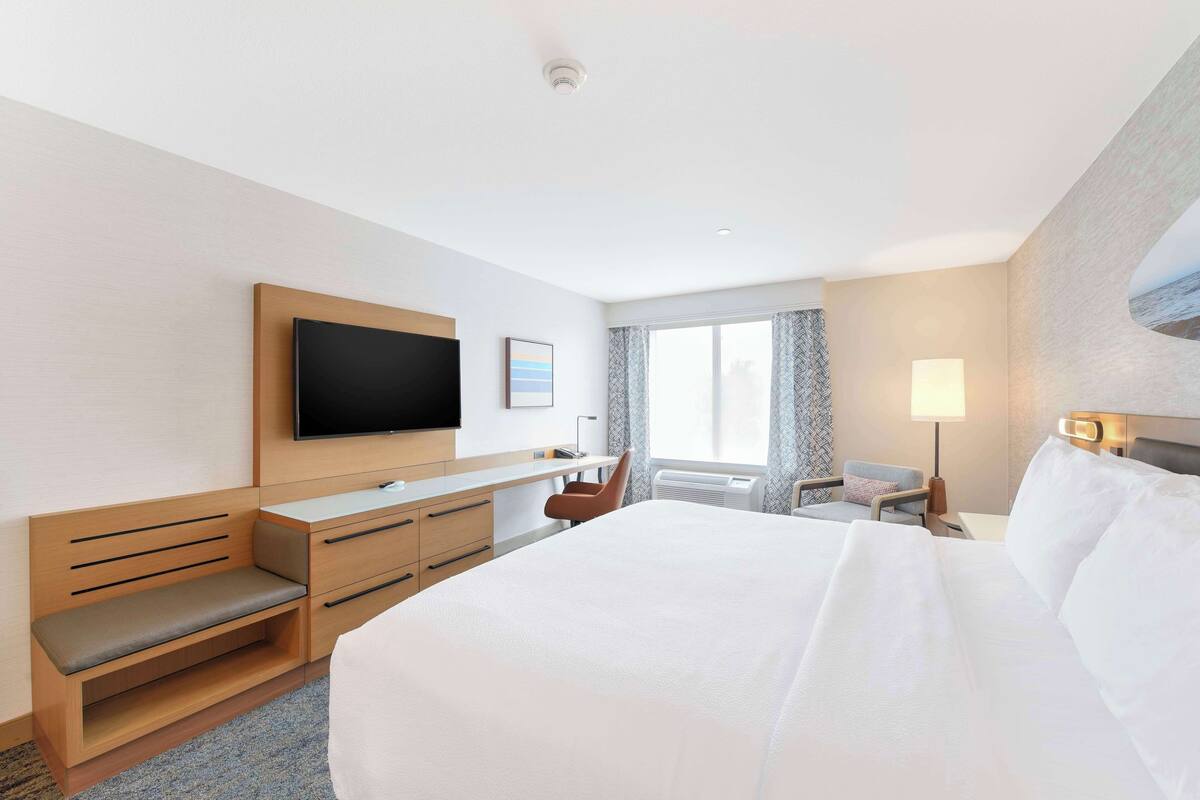 A hotel room with a large bed, a desk with drawers, a flat-screen TV mounted on the wall, a window with curtains, and a bedside table with a lamp.