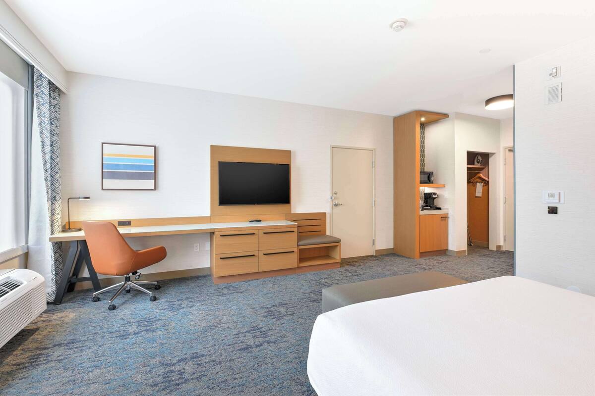 A hotel room with a bed, a desk with a chair, a television, and a kitchenette.