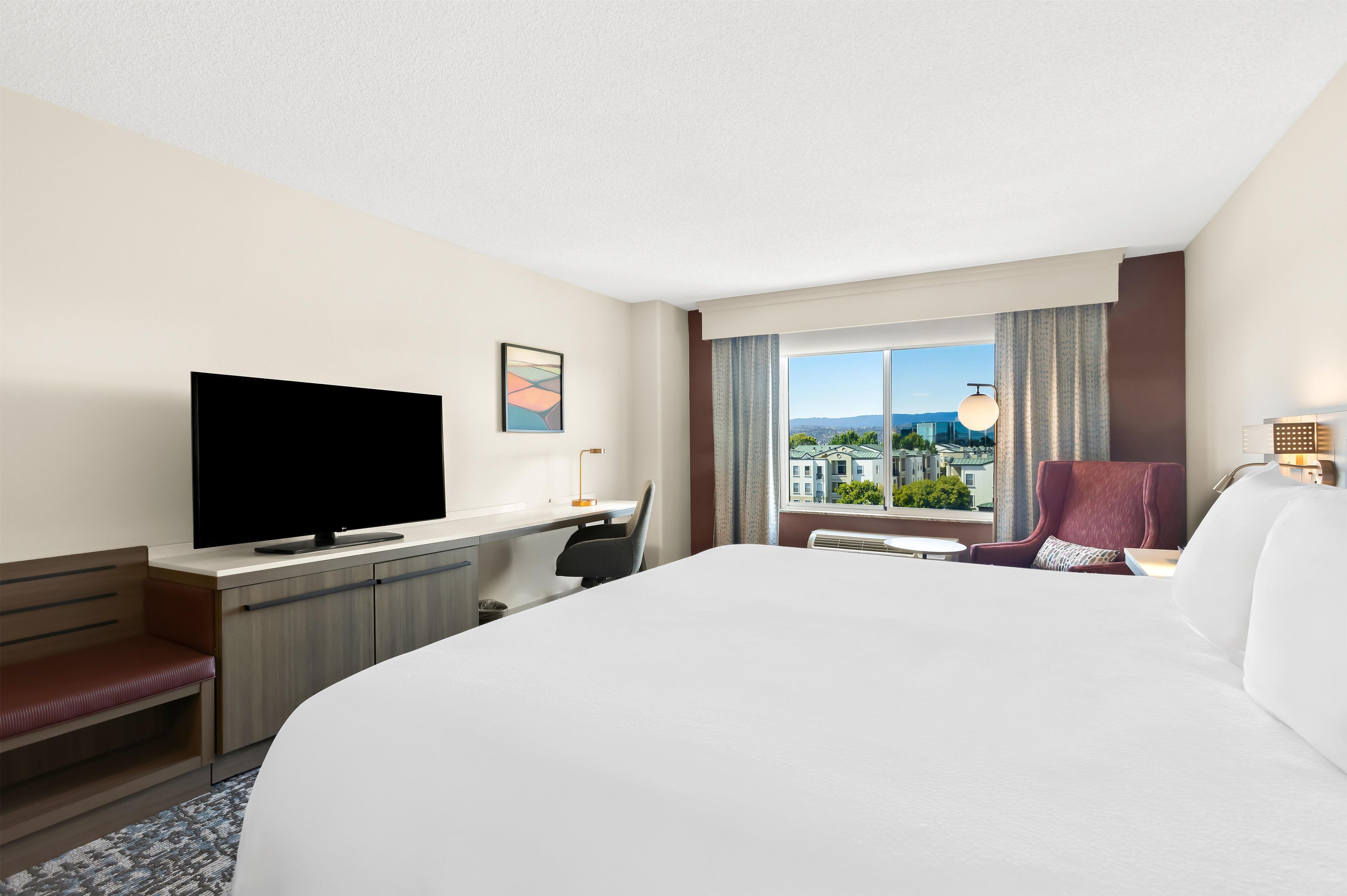 Habitación de hotel con una cama grande, un escritorio, un televisor de pantalla plana, una silla, una ventana con vistas a edificios y una lámpara.