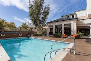 A swimming pool with a metal ladder, surrounded by outdoor furniture and a building with large windows. - Hilton Garden Inn San Mateo (San Mateo)