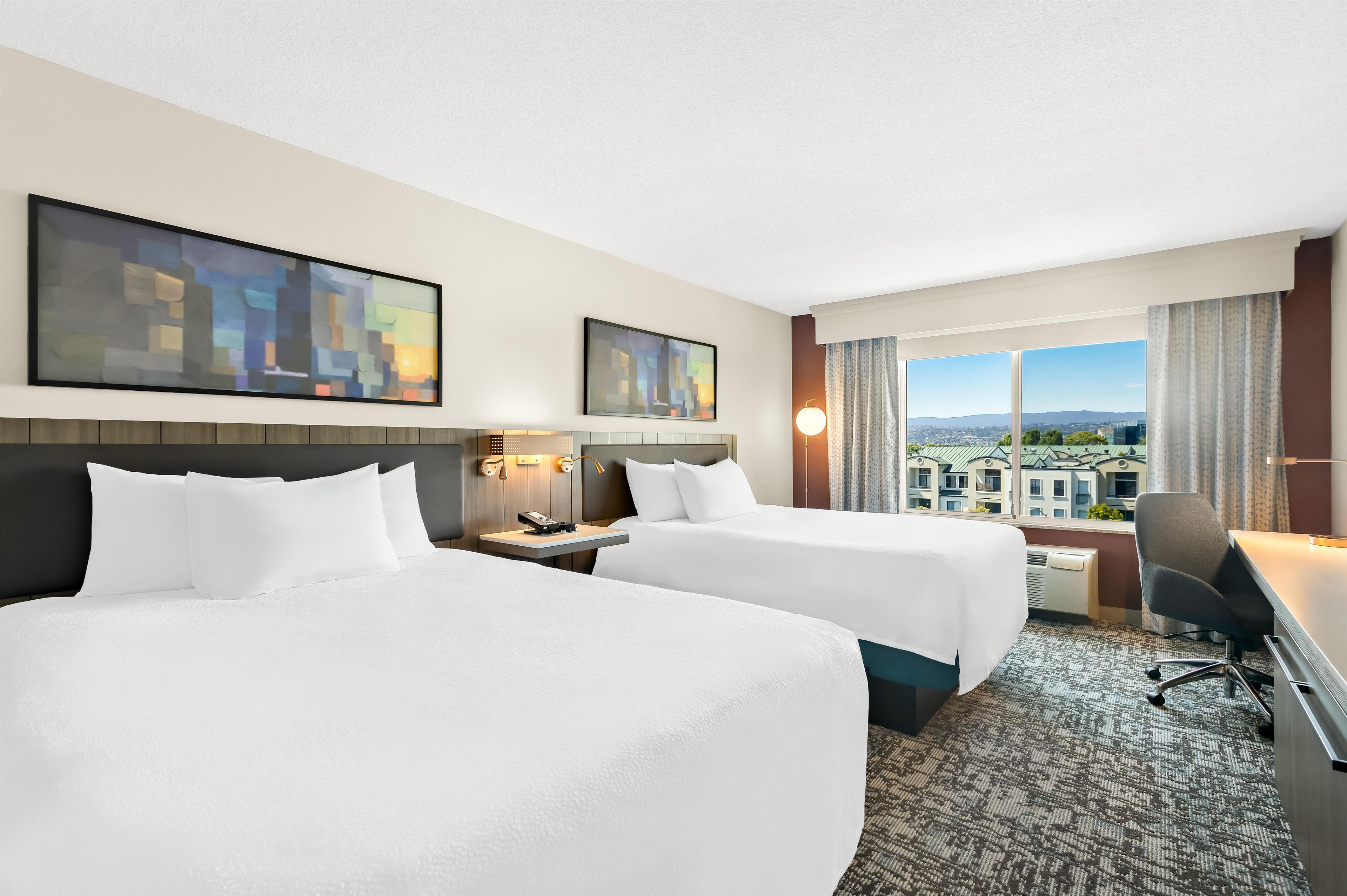 A hotel room with two beds, a desk, a chair, and a view of a cityscape.