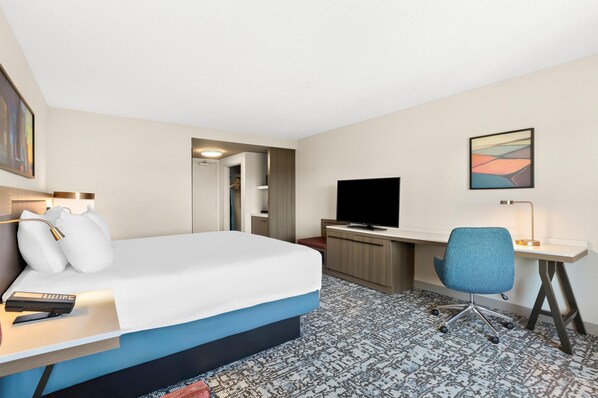 A hotel room with a bed, desk, chair, TV, and artwork.