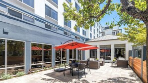 An outdoor patio with tables and chairs, a red umbrella, and a view of a building with large windows. - Hilton Garden Inn San Mateo (San Mateo)
