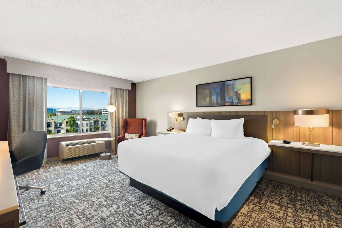 A hotel room with a large bed, a desk, a chair, a window with a view of buildings, and a painting on the wall.