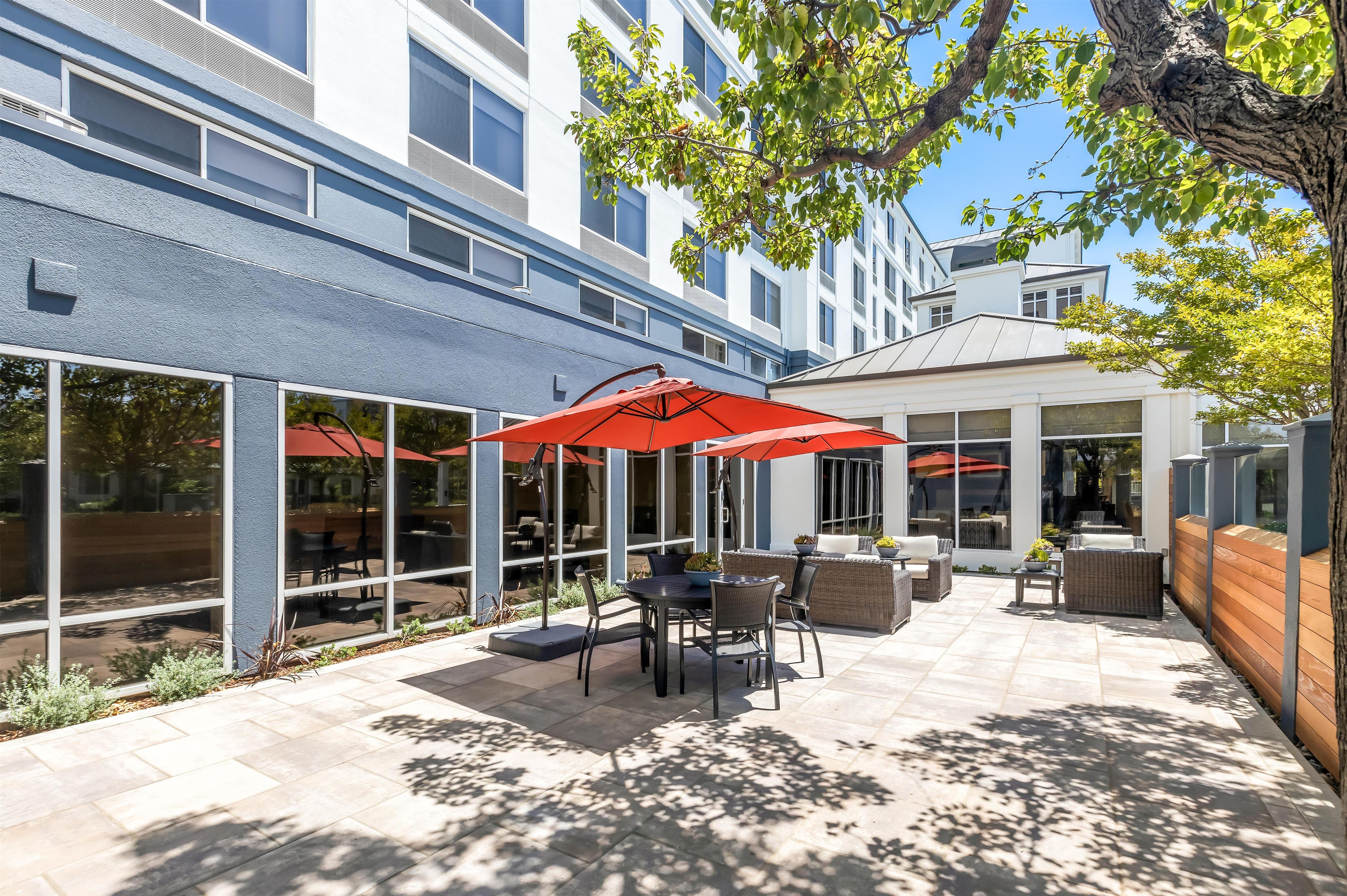 Un área de patio al aire libre con mesas y sillas, sombrillas grandes y vistas a un edificio con ventanas amplias.