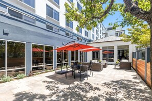 An outdoor patio area with tables and chairs, large umbrellas, and a view of a building with large windows. - Hilton Garden Inn San Mateo (San Mateo)