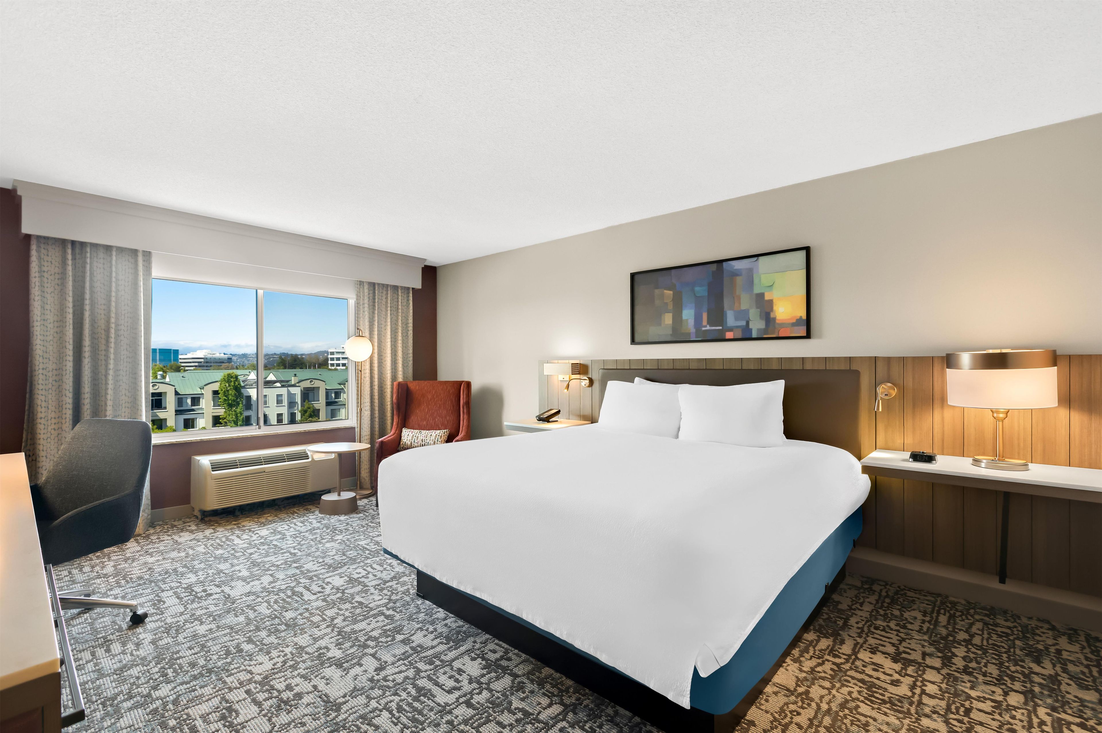 A hotel room with a large bed, a desk, a chair, a window with a view, and a painting on the wall.