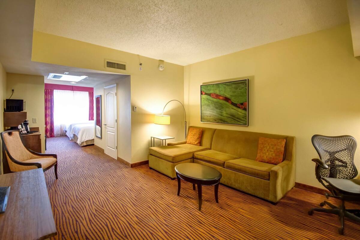Ein Hotelzimmer mit Bett, Sofa, Schreibtisch, Stuhl, Lampe und einem Bild an der Wand.