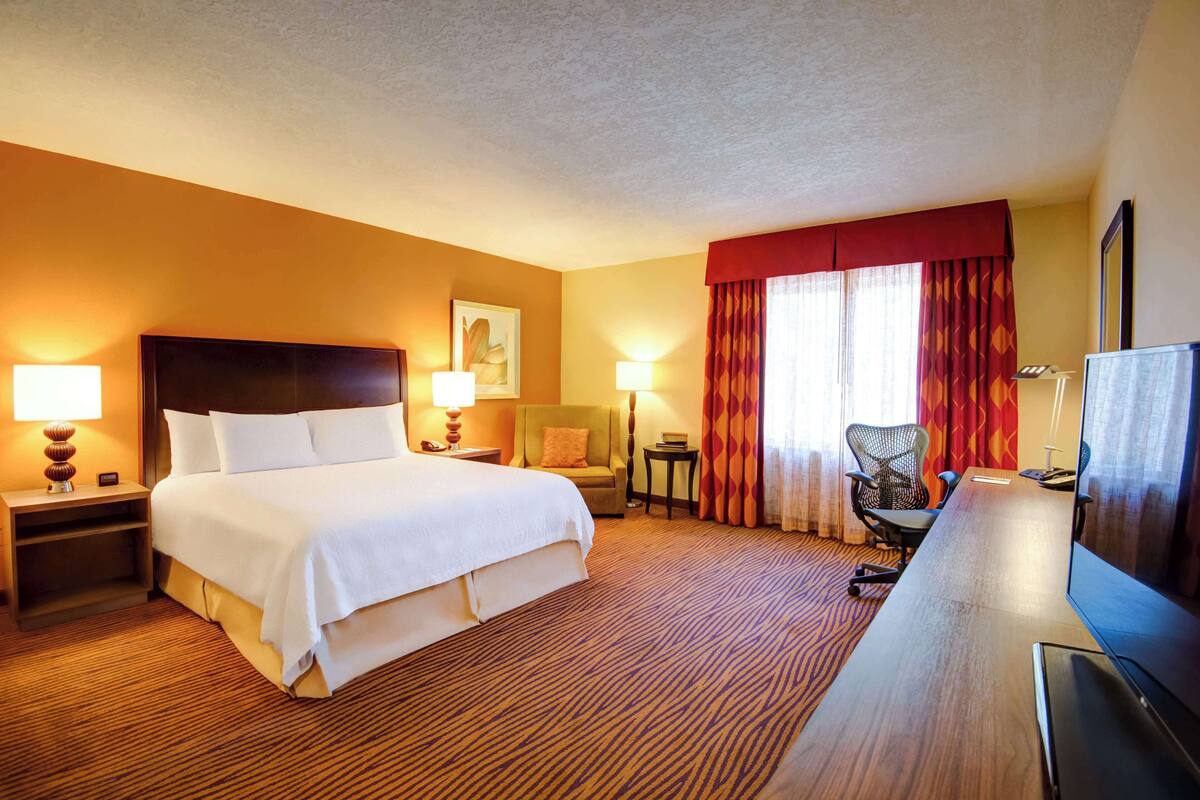 Ein Hotelzimmer mit einem großen Bett, einem Schreibtisch mit Fernseher, einem Sessel und zwei Lampen.
