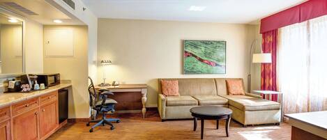 A hotel room with a kitchenette, a sofa, a desk, and a painting on the wall.