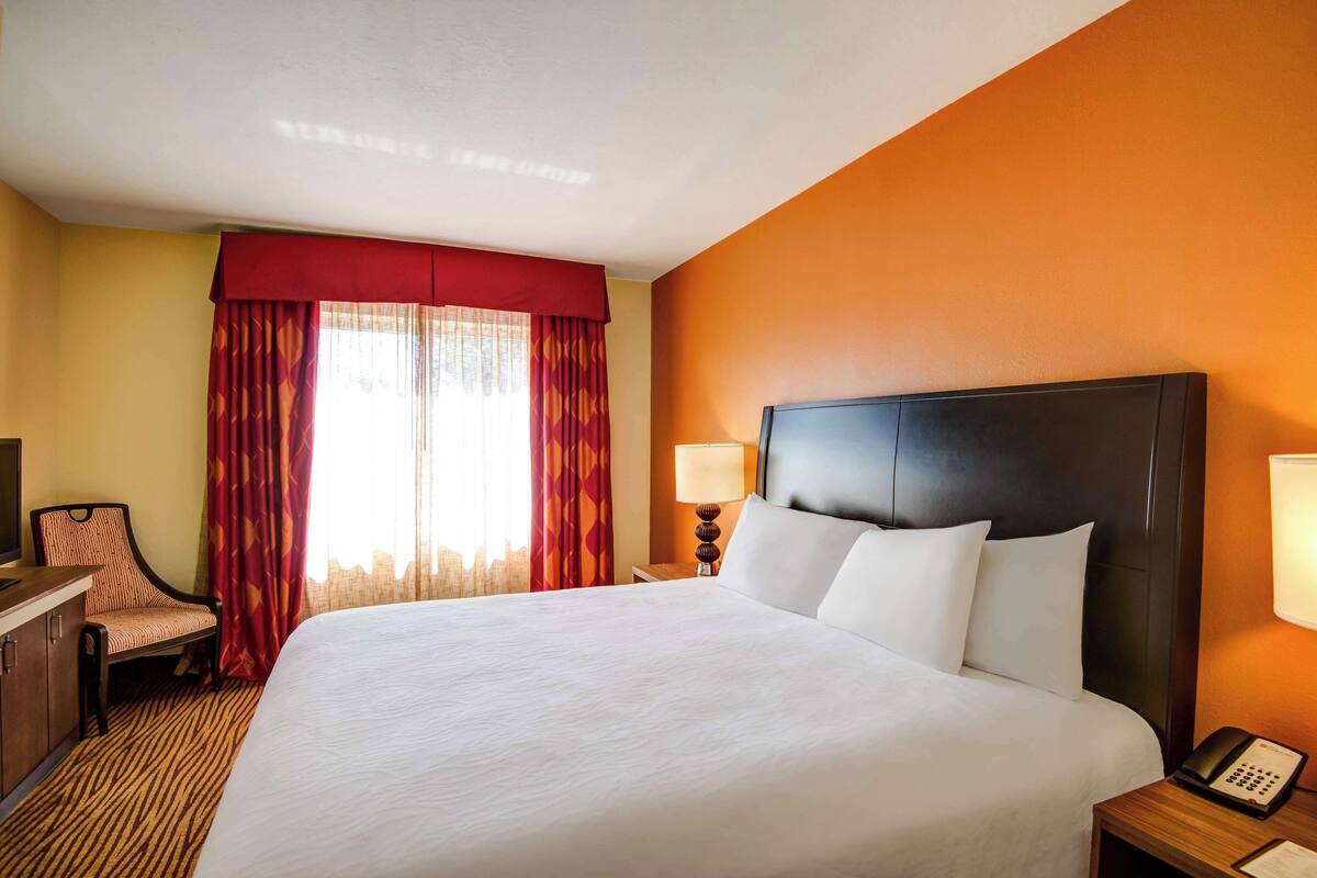 Ein Hotelzimmer mit einem großen Bett, einem Schreibtisch mit Stuhl, einem Telefon und einem Fenster mit Vorhängen.
