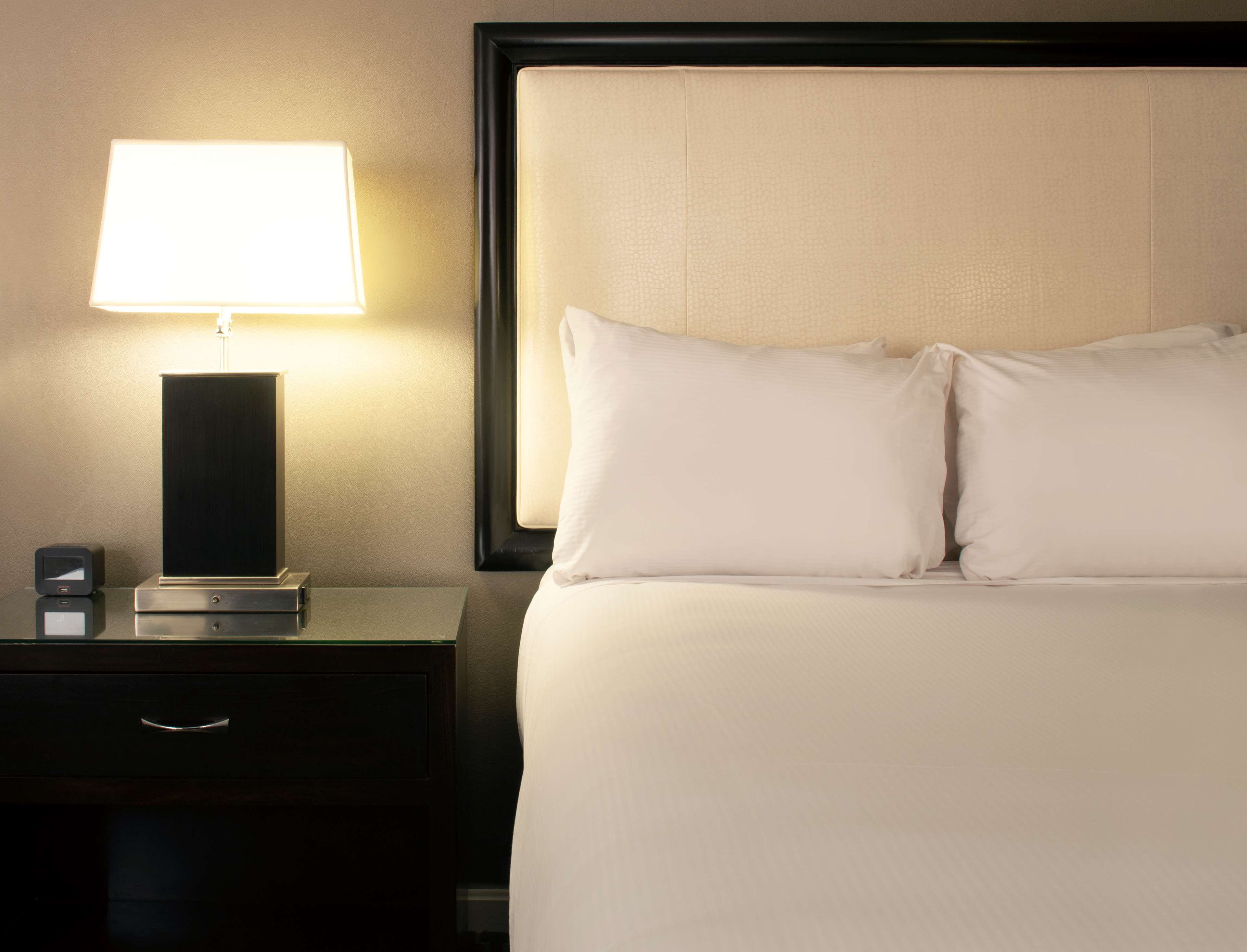 A neatly made bed with white linens and pillows, a bedside table with a lamp, and a small electronic device.