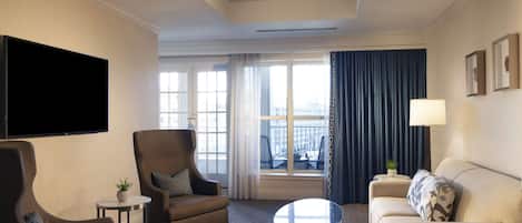 A modern hotel room with a flat-screen TV, a sofa, a chair, a round coffee table, and a large window with curtains.