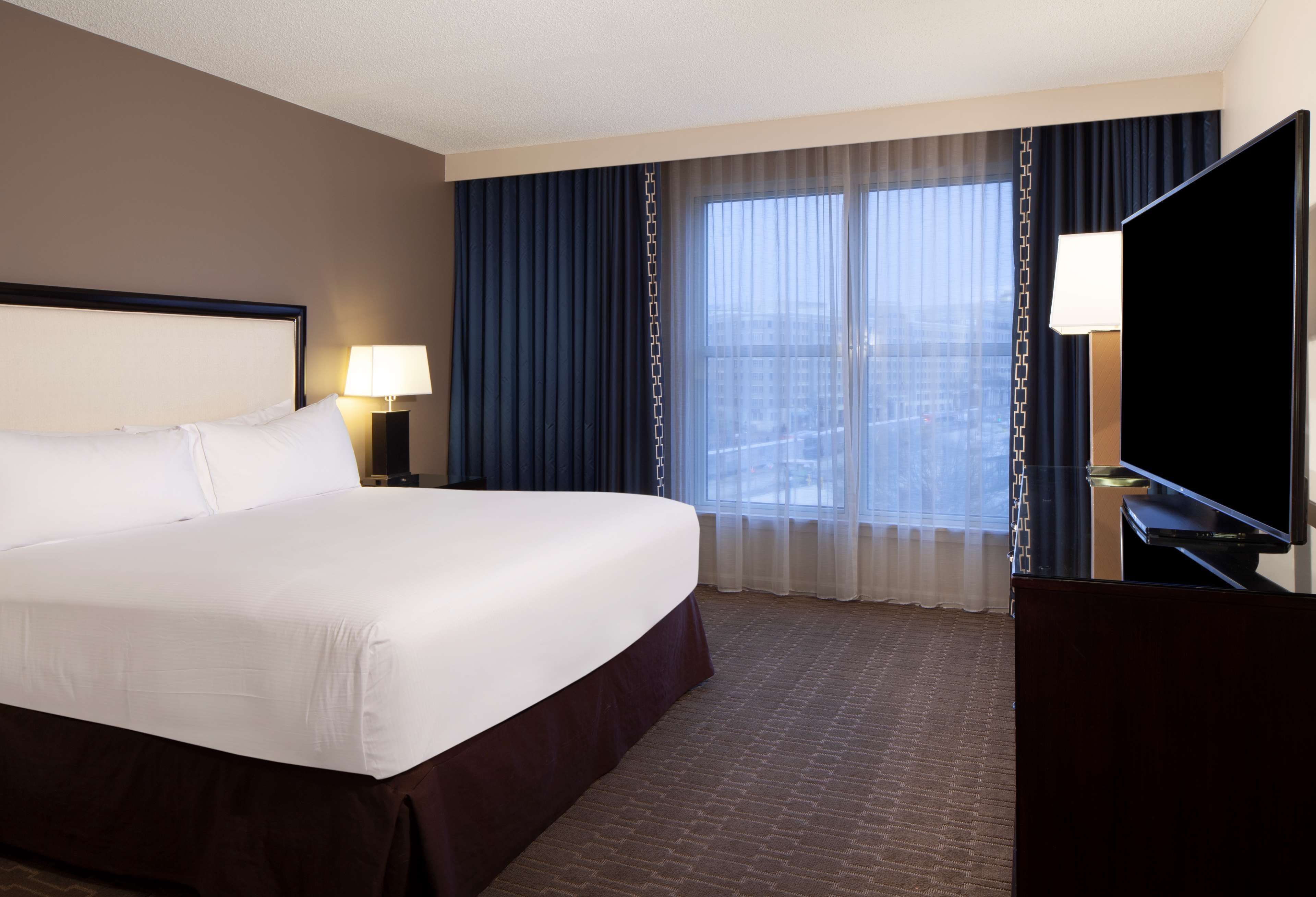 Een hotelkamer met een groot bed, een bureau met een flatscreen televisie, een lamp en uitzicht op de stad door grote ramen.