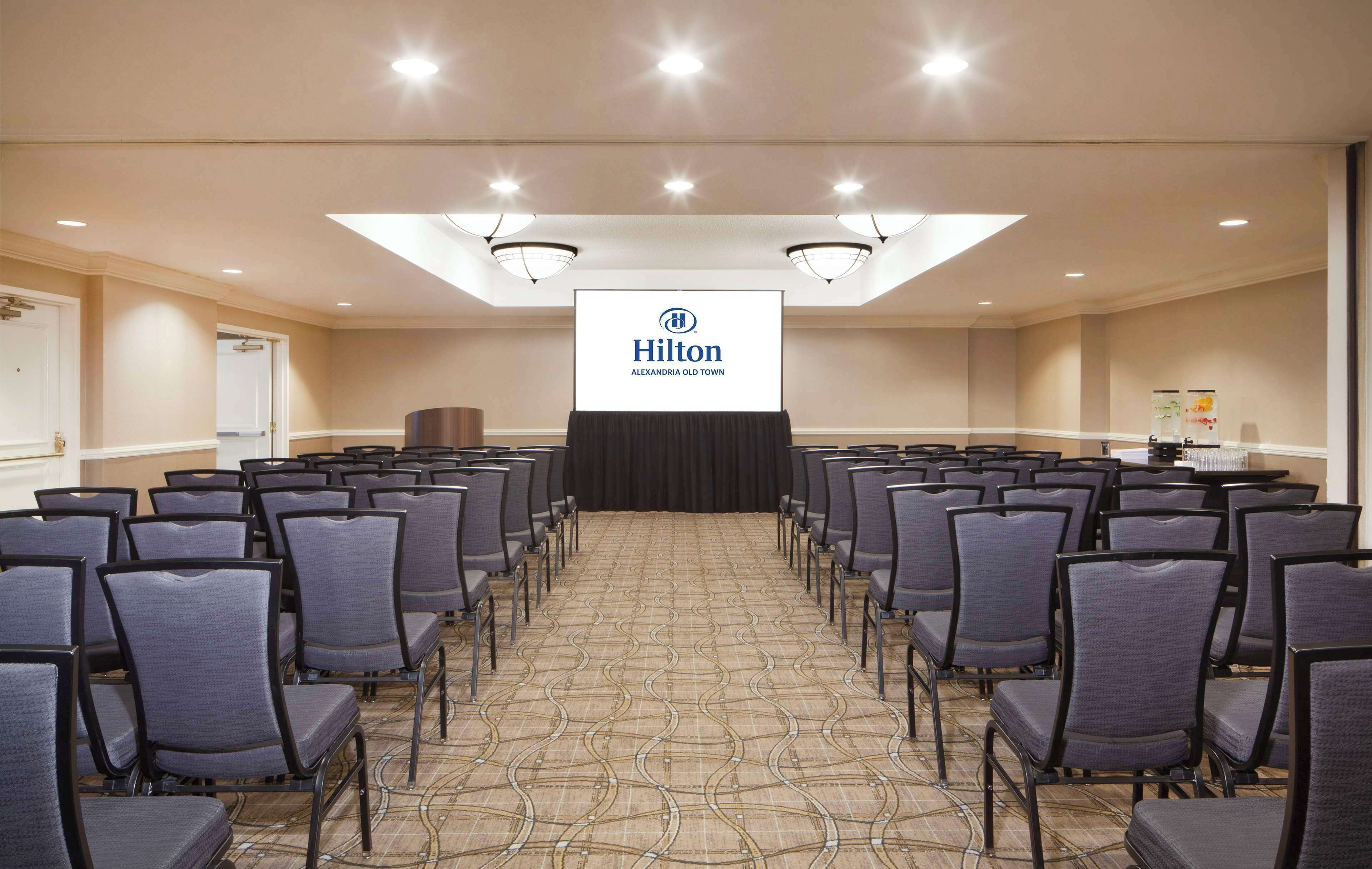 A conference room at Hilton Alexandria Old Town with rows of chairs, a podium, and a large screen displaying the Hilton logo.