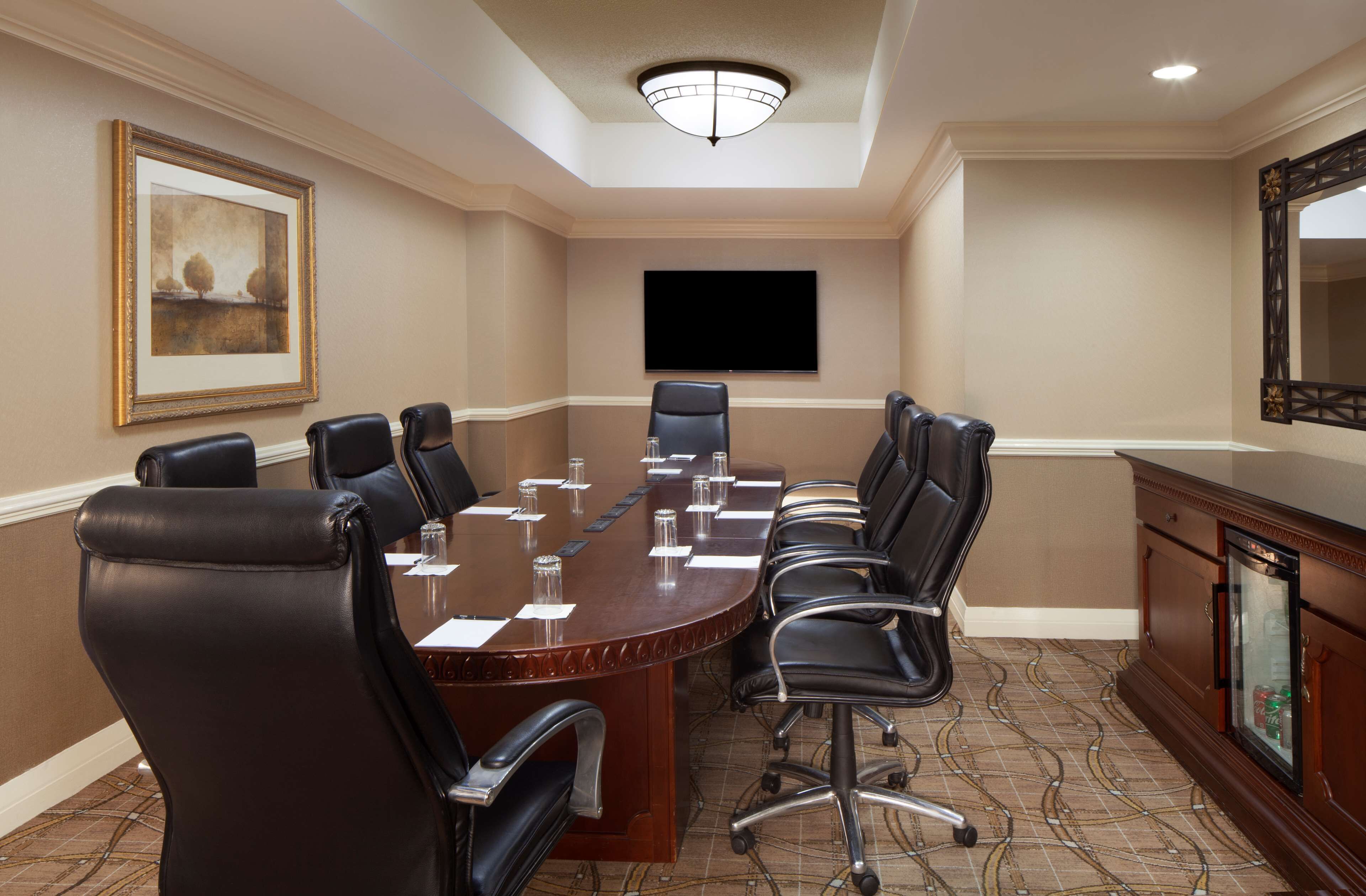 A conference room with a large oval table, black leather chairs, a flat-screen TV, and a framed picture on the wall.