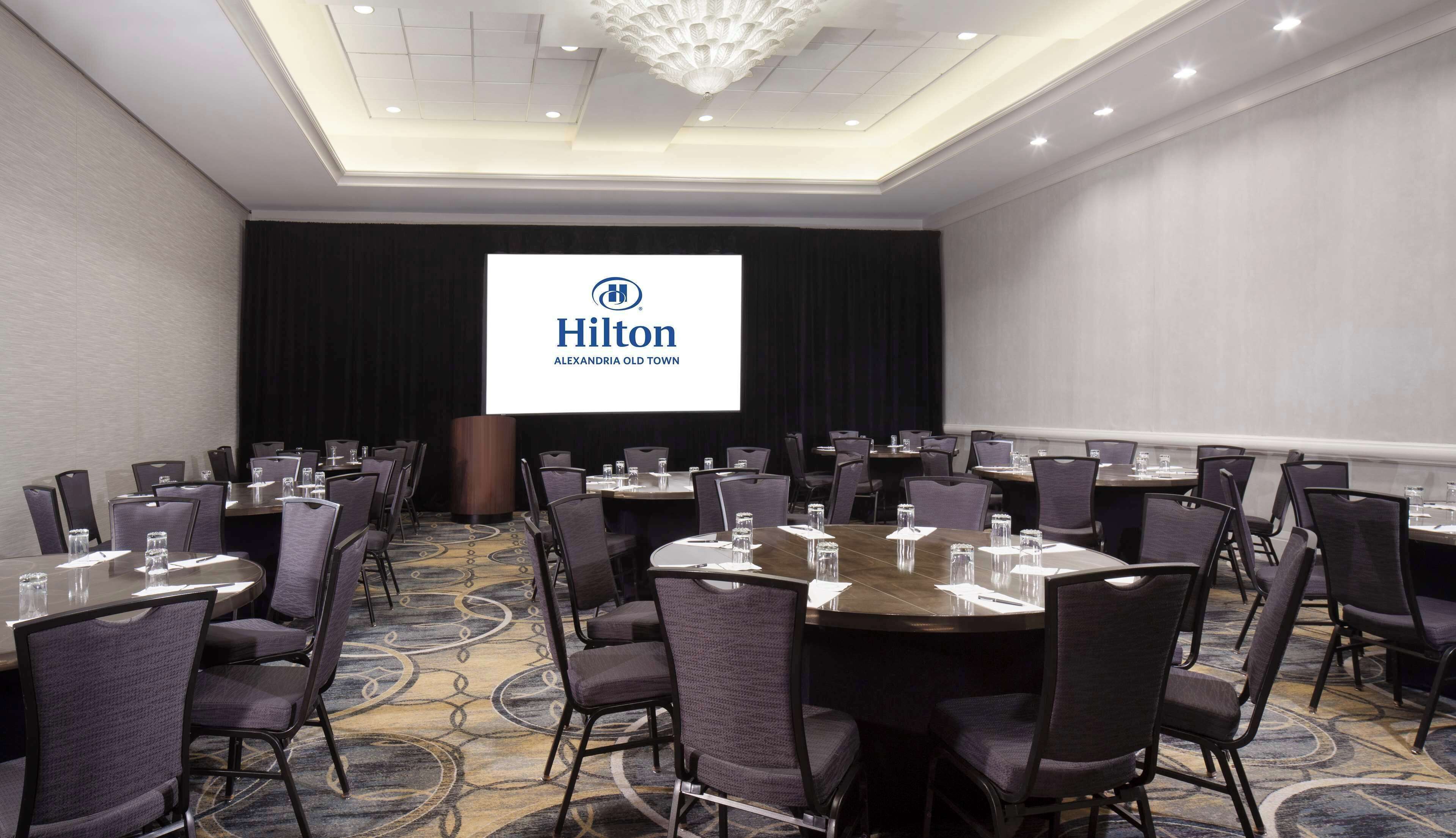 Une salle de conférence dotée d’un grand écran affichant le logo de Hilton, de tables disposées avec des chaises et d’un sol recouvert de moquette.