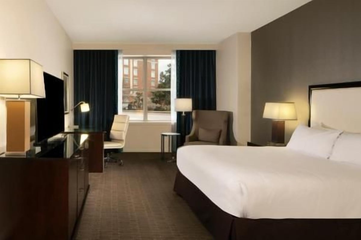 A hotel room with a bed, two chairs, a desk, a lamp, and a window with curtains.