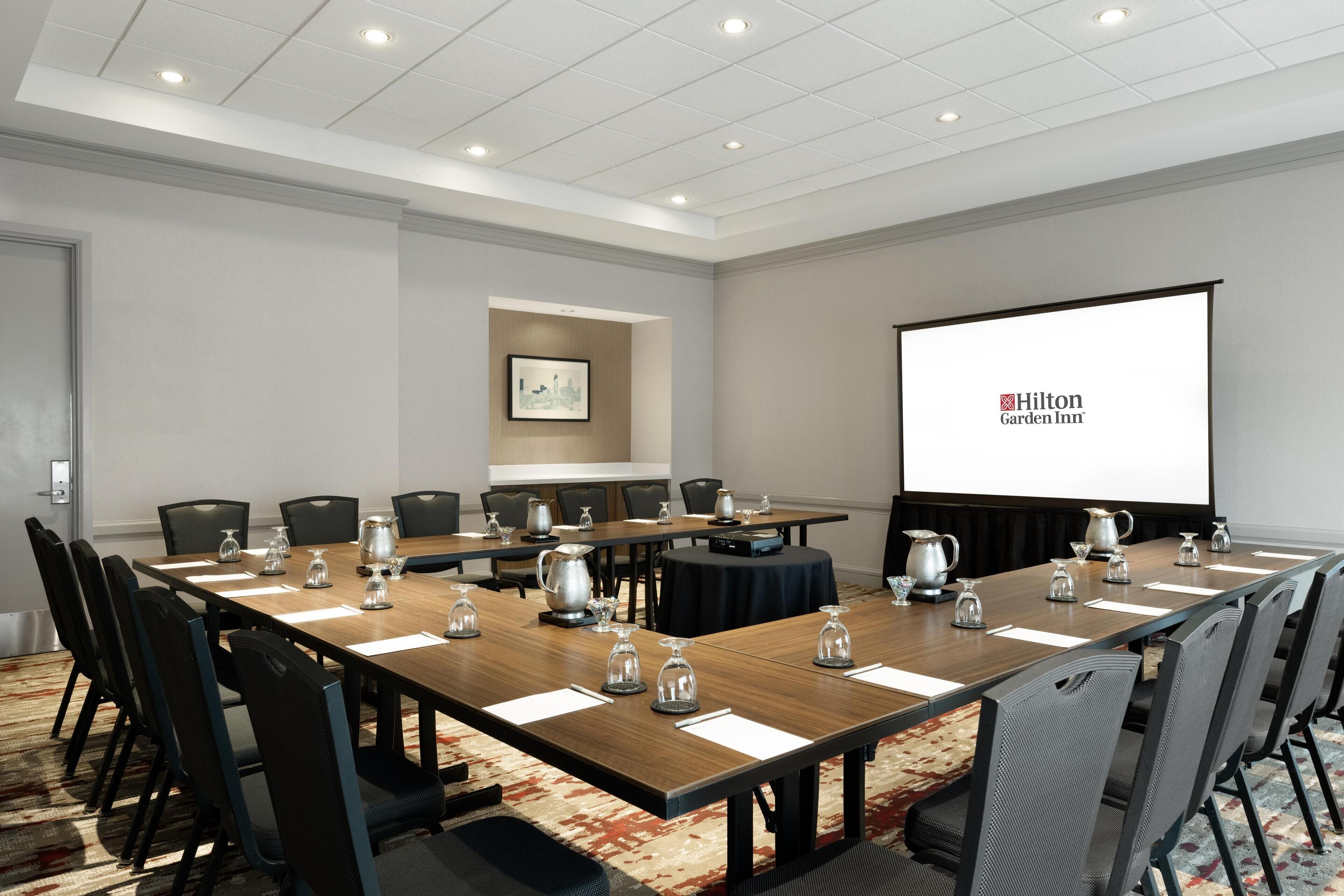 Ein Konferenzraum mit einem langen Tisch, Stühlen, einer Projektionsleinwand, auf der das Hilton Garden Inn Logo zu sehen ist, und einem gerahmten Bild an der Wand.