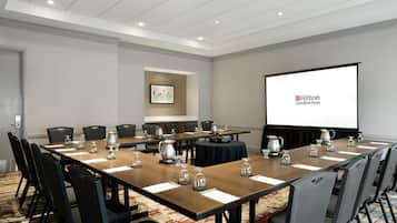 Ein Konferenzraum mit einem langen Tisch, Stühlen, einer Projektionsleinwand, auf der das Hilton Garden Inn Logo zu sehen ist, und einem gerahmten Bild an der Wand.