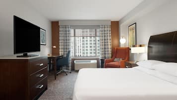A hotel room with a large bed, a desk with a chair, a television, a lamp, and a window with curtains.