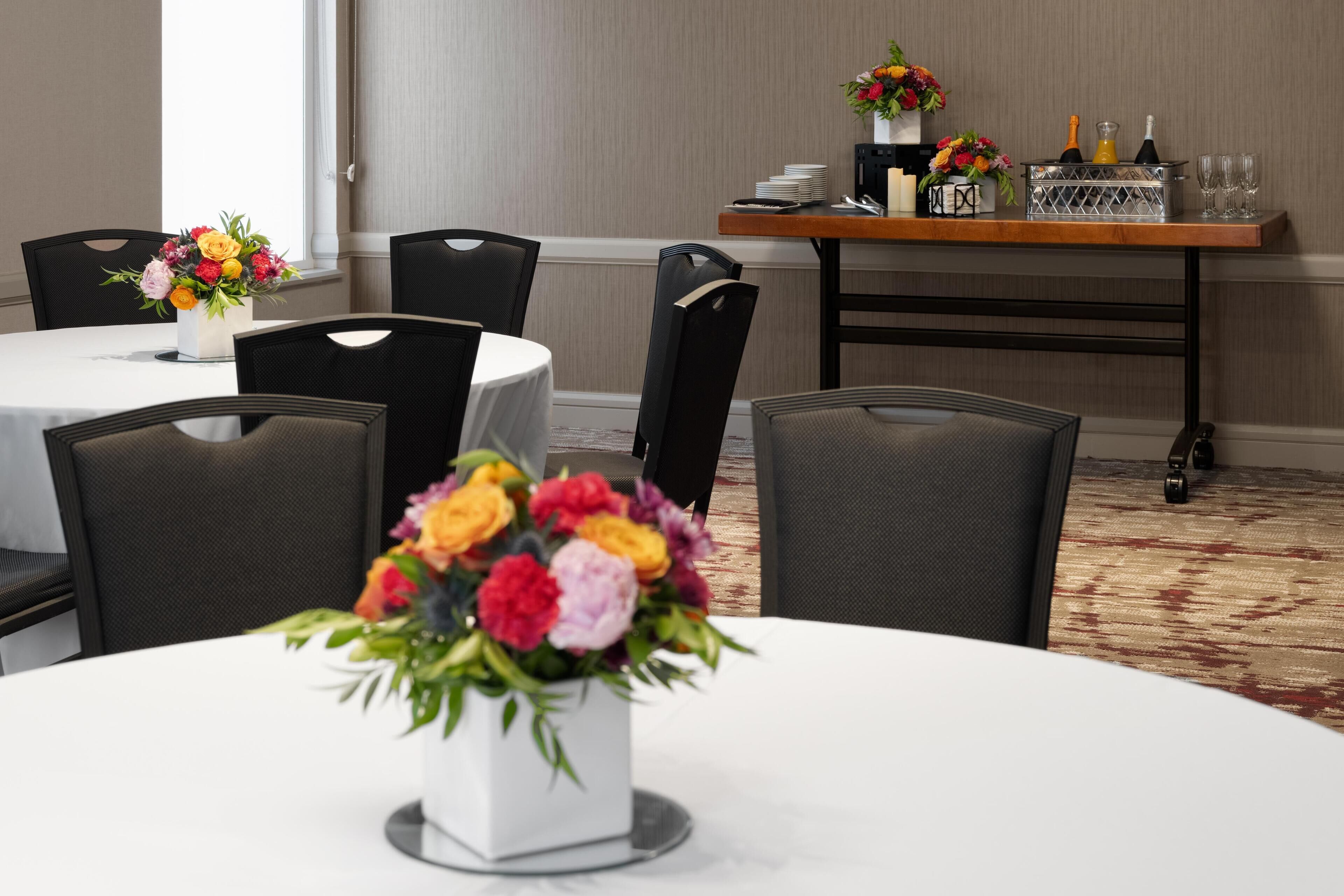 A dining area with round tables, black chairs, and floral centerpieces. There is a bar area with a tray table and glasses.