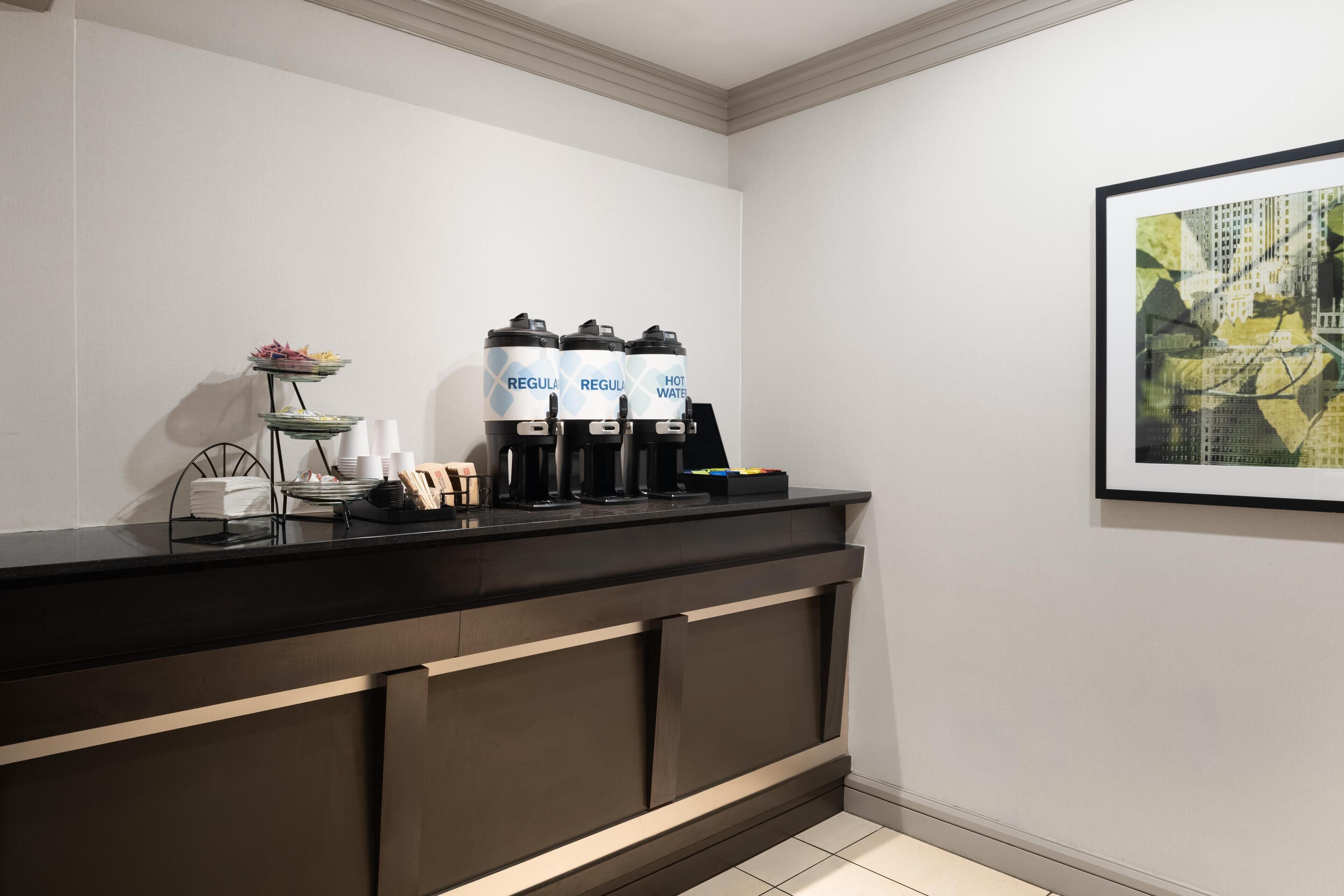 A modern hotel reception area with a dark counter, a coffee machine, and water dispensers labeled 'REGULA HOT WATER'.