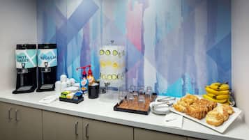 A breakfast bar with coffee makers, a juice dispenser, and a tray of pastries and bananas.