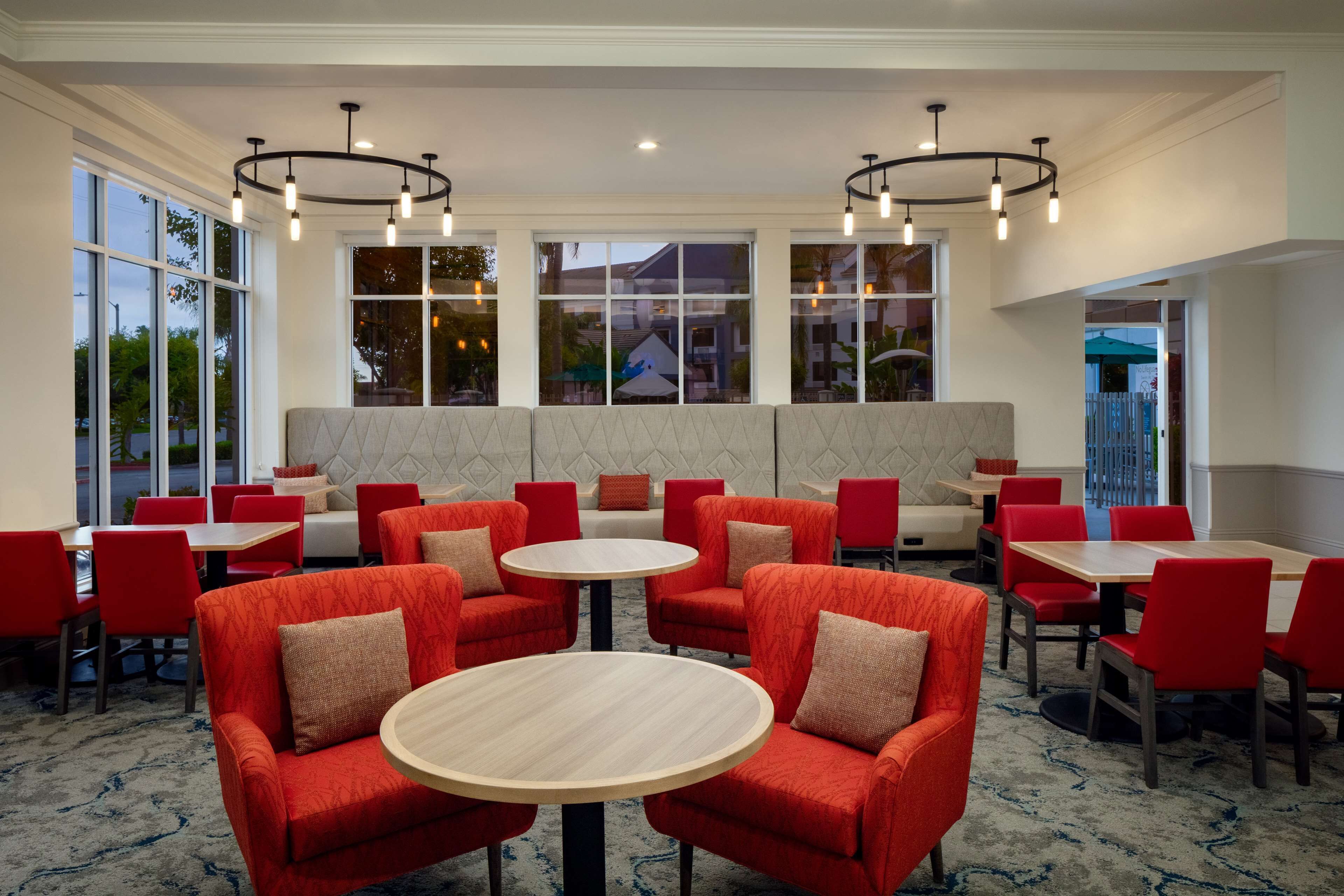 Un área de comedor moderna con sillas tapizadas en rojo y mesas redondas, ventanas amplias con vistas y lámparas colgantes.
