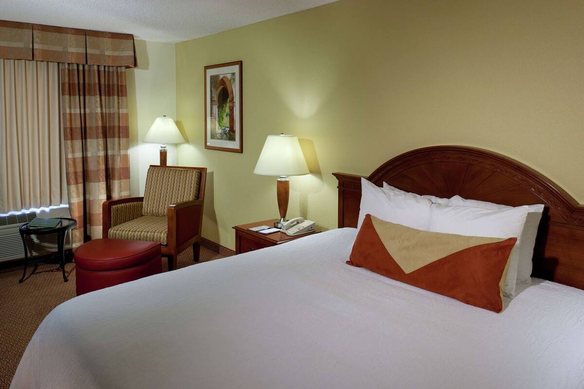 Una habitación de hotel con cama, una silla, un escritorio, una lámpara y un cuadro en la pared.