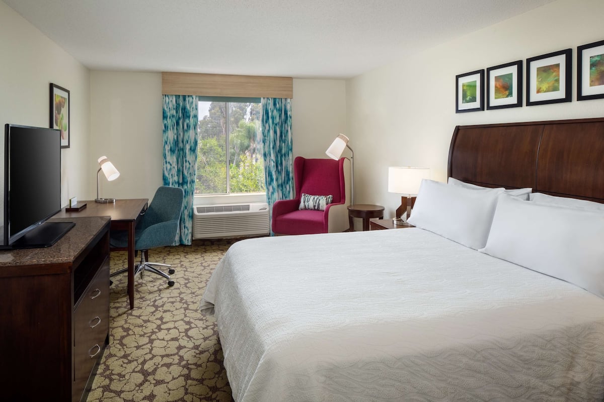 Habitación de hotel con una cama grande, un escritorio con computadora, una silla, un televisor y una ventana con cortinas.